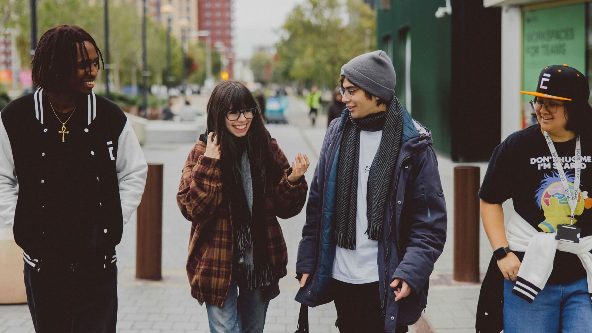 A group of four friends walking and chatting on a city street, dressed in casual winter clothing and appearing happy and relaxed.