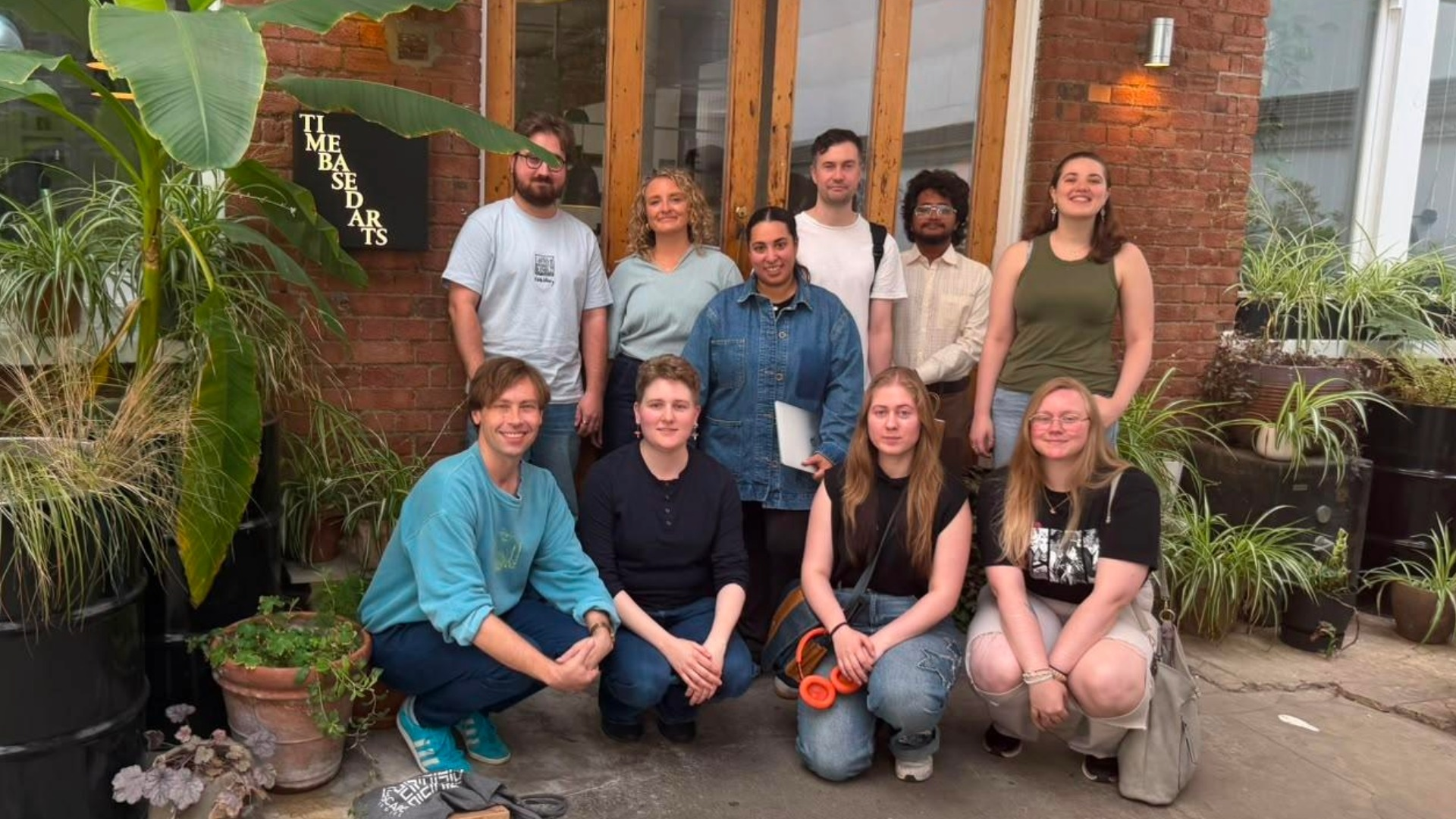 A group of ten people posing together in front of a building with plants and a sign reading "Time Based Arts" on a brick wall.