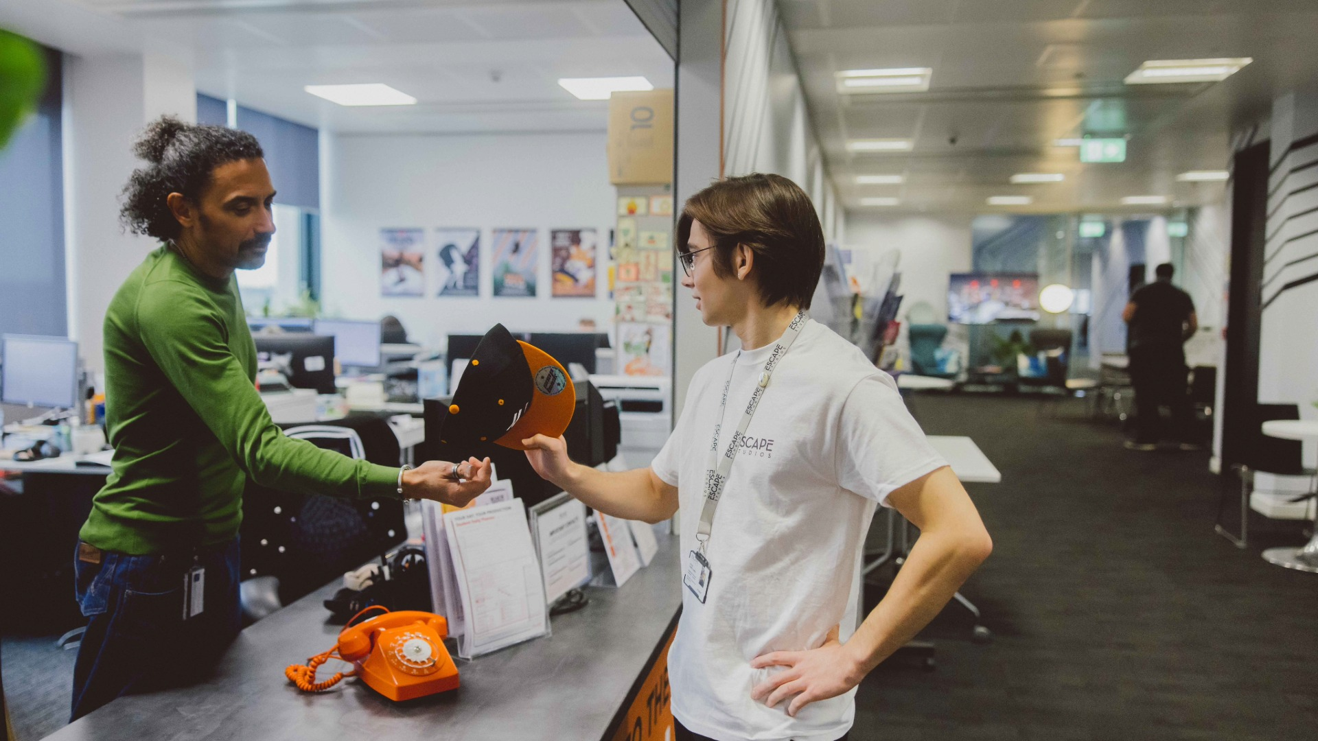Two people in an office; one hands a black and orange cap to the other at a desk with an orange rotary phone.